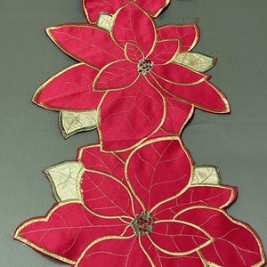 Puede incluir: Un camino de mesa rojo con flores de pascua y adornos dorados. El camino de mesa presenta múltiples diseños de flores de pascua con costuras detalladas y un tema festivo. Las hojas son de color verde claro.