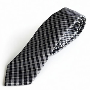 May include: A black and white chequered tie, folded on a white surface. The tie has a repeating pattern of small squares in shades of black, grey, and white. A classic accessory for formal or business attire.