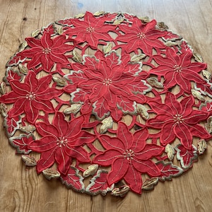 May include: A circular red poinsettia placemat with gold embroidered detailing. The placemat has a cut-out design featuring multiple poinsettia flowers and gold-coloured leaves. The decorative item is placed on a wooden surface.