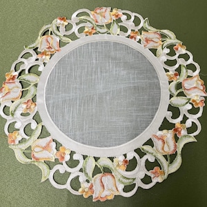 Könnte beinhalten: Ein weißer Tischläufer aus Spitze mit einem Blumendesign mit orangefarbenen und grünen Tulpen. Der Läufer hat eine runde Mitte und einen gewellten Rand.