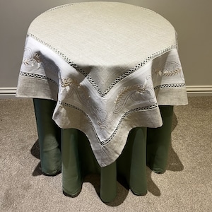 Puede incluir: Un mantel de lino blanco con un diseño floral bordado. El mantel está drapeado sobre una mesa verde.