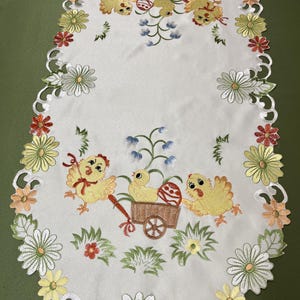 May include: White oval table runner with embroidered yellow chicks pulling a wagon with an Easter egg. The runner has a scalloped edge with embroidered flowers in shades of yellow, green, red, and orange.