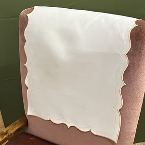 Puede incluir: Funda blanca para el respaldo de la silla con borde festoneado y bordado dorado. La funda está colocada en una silla de terciopelo rosa.