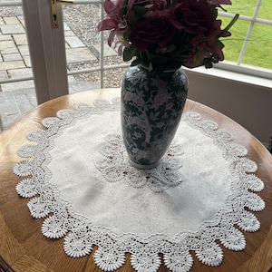 Könnte beinhalten: Ein runder Holztisch mit einer weißen Spitzendecke und einer dekorativen Vase mit rosa und burgunderfarbenen Blumen. Die Vase ist dunkelgrün mit einem floralen Muster.