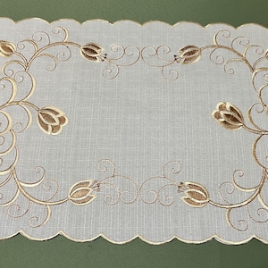 Può includere: Un sottobicchiere rettangolare bianco sporco con bordo smerlato. Presenta un disegno ricamato di fiori marroni e viticci. Un elemento decorativo per la tavola.