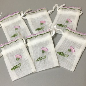 May include: Six small white fabric drawstring bags with green and purple embroidered thistles. The bags have a scalloped edge and are tied with white ribbon.