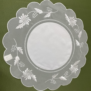 May include: A white, scalloped-edge doily with embroidered floral and leaf patterns. The center is a solid white circle, surrounded by a detailed, stitched border. The delicate design adds a touch of elegance.