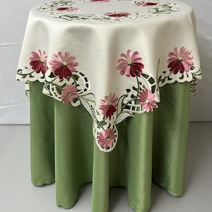 Puede incluir: Mantel de mesa blanco con flores bordadas de color rosa y rojo y un borde festoneado. El mantel está sobre una mesa verde.