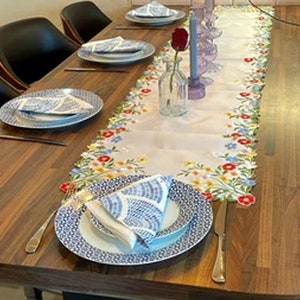 May include: A wooden dining table set for a meal. The table is adorned with a floral embroidered table runner, blue and white patterned plates, and folded napkins. A red rose in a glass vase and candles add to the decor.