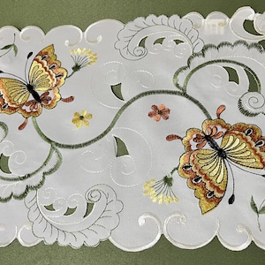 May include: White embroidered table runner with a scalloped edge featuring two butterflies with brown and yellow wings and green foliage.