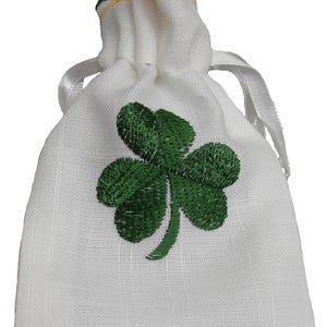 May include: A white linen drawstring bag featuring a green embroidered shamrock. The top edge is trimmed with green and gold detailing. A white satin ribbon is used to close the bag. The bag is a symbol of St. Patrick's Day.