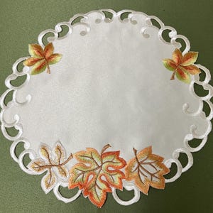 May include: White round embroidered table runner with a scalloped edge and a design of autumn leaves in shades of orange, yellow, and green.