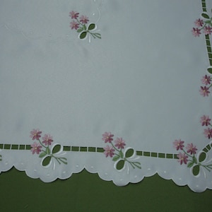 May include: White embroidered tablecloth with a scalloped edge. The tablecloth features a delicate floral design with pink flowers and green leaves. The flowers are embroidered along the border and in the corners.