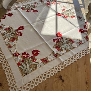 Pode incluir: Uma toalha de mesa branca com uma delicada borda de renda e um desenho floral bordado com flores vermelhas, folhas verdes e margaridas brancas.