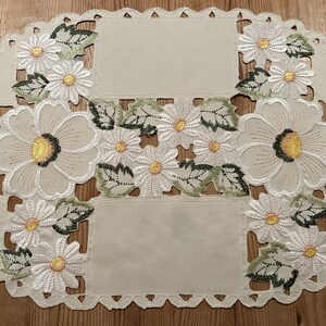 Può includere: Un runner da tavolo ovale bianco con un delicato disegno floreale. Il runner presenta margherite ricamate con foglie verdi e un bordo smerlato.