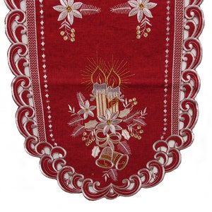 Puede incluir: Camino de mesa rojo con borde de encaje blanco y diseño navideño bordado. El diseño presenta tres velas, acebo y campanas.