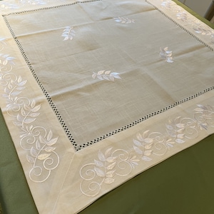 Puede incluir: Mantel de mesa de lino blanco con un borde bordado de hojas y espirales blancas. El mantel tiene una textura sutil y es perfecto para una cena formal.