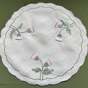 May include: A white, round doily with a scalloped edge, embroidered with thistle flowers. The embroidery uses purple, green, and grey threads. The fabric has a subtle plaid pattern. The doily is a traditional decorative item.