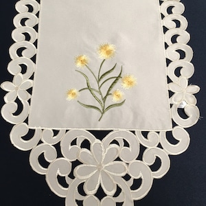 May include: White table runner with a delicate floral embroidery design. The runner features a cluster of yellow flowers with green stems and leaves. The edges of the runner are adorned with a scalloped lace pattern.