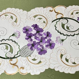 Puede incluir: Un camino de mesa blanco con un delicado diseño floral. El camino de mesa presenta un patrón repetitivo de flores moradas y hojas verdes sobre un fondo blanco con detalles dorados.