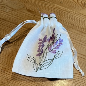 May include: A white drawstring bag with a lavender flower design embroidered on the front. The bag has a white drawstring closure.