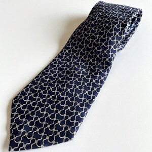 Op de afbeelding: Marineblauwe stropdas met een herhalend wit, abstract patroon. De stropdas is gemaakt van een gladde stof en wordt gepresenteerd op een witte achtergrond. Een klassiek accessoire voor formele of zakelijke kleding.