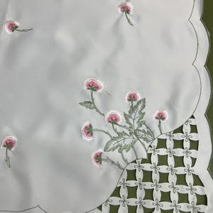 May include: White tablecloth with embroidered pink and white flowers and green leaves. The edge is scalloped and outlined in green. A cut-out section features a geometric design.