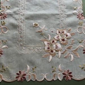 May include: A rectangular table runner made of sheer, light-colored fabric featuring intricate embroidery. The design includes scalloped edges, floral motifs in shades of dusty rose, brown, and cream, and a decorative stitched border pattern.