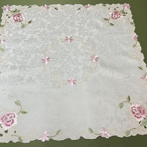 以下が含まれることがあります： ピンクの花柄が繊細に刺繍された白いテーブルクロス。テーブルクロスは波打つ縁取りになっており、ピンクのバラと葉の繰り返し模様が特徴です。