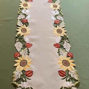May include: A beige table runner featuring embroidered sunflowers, white daisies, and red buds. The floral design is bordered by green and beige leaves. The runner is on a green surface.
