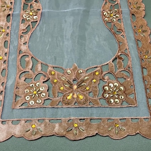 May include: A decorative table runner with a light blue sheer center and a brown, scalloped border. The border features cut-out floral and butterfly designs, embellished with gold and yellow sequins. A delicate home decor item.
