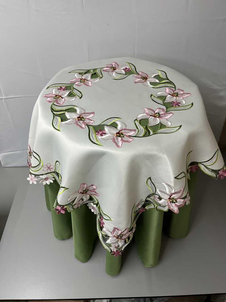 May include: White round tablecloth with embroidered pink and green lilies around the edge. The tablecloth is draped over a green table.