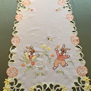 Può includere: Runner da tavola pasquale color crema con ricami di coniglietti, uova di Pasqua, fiori e una farfalla. I bordi sono smerlati con ritagli floreali gialli. Il runner è lungo circa 152 cm.