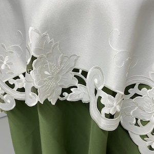May include: White tablecloth with a delicate floral embroidery design. The embroidery features intricate cutouts and a raised floral pattern. The tablecloth is shown draped over a green surface.