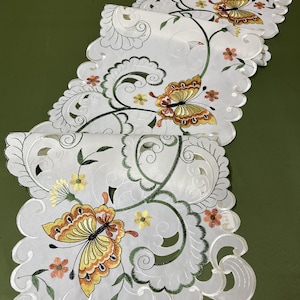 Puede incluir: Caminero de mesa blanco bordado con un borde festoneado y un patrón repetido de hojas verdes, mariposas naranjas y amarillas y pequeñas flores naranjas y amarillas.