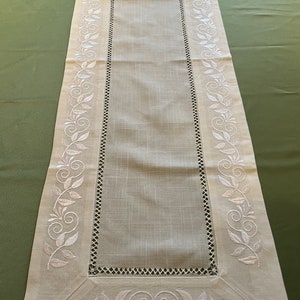 Könnte beinhalten: Ein weißer Tischläufer aus Leinen mit einem gestickten Blumendesign. Das Design zeigt weiße Blätter und Spiralen entlang der Kanten des Läufers.