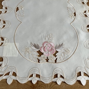 Könnte beinhalten: Weißer bestickter Tischläufer mit einem zarten Blumendesign mit einer rosa Rose in der Mitte. Der Läufer hat einen gewellten Rand und ein spitzendes Muster.