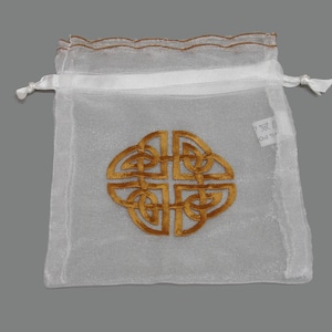 May include: A white, sheer drawstring bag featuring a gold Celtic knot design embroidered on the front. The bag has a gold trim along the top and white drawstrings.