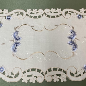 May include: Cream-coloured rectangular table runner with intricate floral embroidery and cut-out edges. Features blue and gold floral accents. Decorative home decor item.