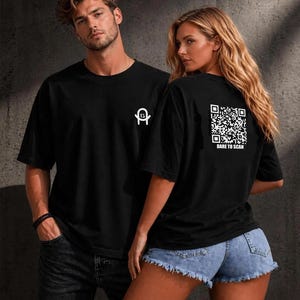 Può includere: Una t-shirt nera a maniche corte con un piccolo logo bianco sul petto e una grande grafica con codice QR bianco sul retro, accompagnata dalla scritta DARE TO SCAN stampata sotto il codice. Il capo presenta un girocollo e una vestibilità ampia.