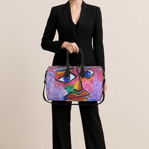 May include: A colorful duffel bag featuring an abstract face design with blue eyes, red lips, and yellow and green accents. The bag has black handles and a shoulder strap. The bag is held by a person wearing a black suit.