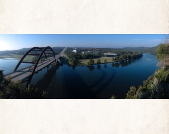 May include: An aerial view of a red steel bridge over a blue river. The bridge has two lanes of traffic and is surrounded by green trees. There are buildings in the distance.