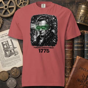 May include: A red t-shirt featuring a graphic of a man with green-tinted glasses and the text "GIVE ME LIBERTY OR GIVE ME DEATH." The shirt also includes the words "SYSTEM MANGRANT" and "FORCED EXECUTABLE: 1775."