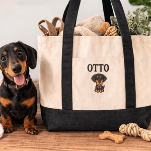 May include: A black and tan dachshund dog sits next to a cream and black tote bag with the name "OTTO" and a dachshund graphic. The bag contains dog toys and is next to a leash and a bone-shaped treat.