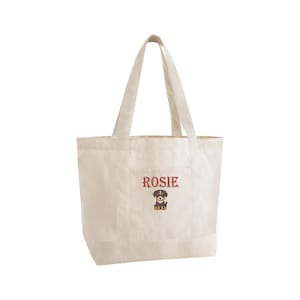 May include: Off-white canvas tote bag with a pocket. The word "Rosie" is embroidered in burgundy above a cartoon dog illustration. The bag has two handles for carrying and is designed for everyday use.