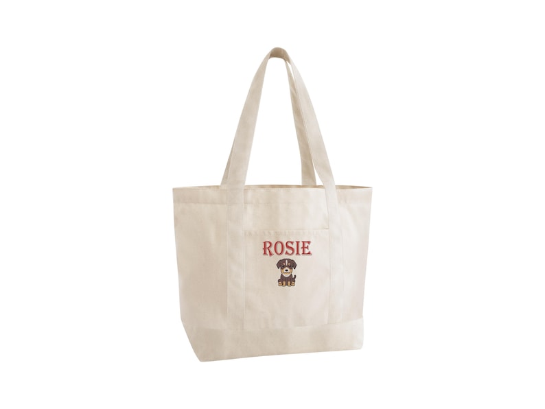 May include: Off-white canvas tote bag with a pocket. The word "Rosie" is embroidered in burgundy above a cartoon dog illustration. The bag has two handles for carrying and is designed for everyday use.