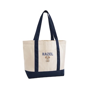 May include: Cream-colored tote bag with navy blue accents. The bag features navy blue handles and a navy blue bottom. The word "HAZEL" is embroidered in navy blue, along with a small puppy graphic.
