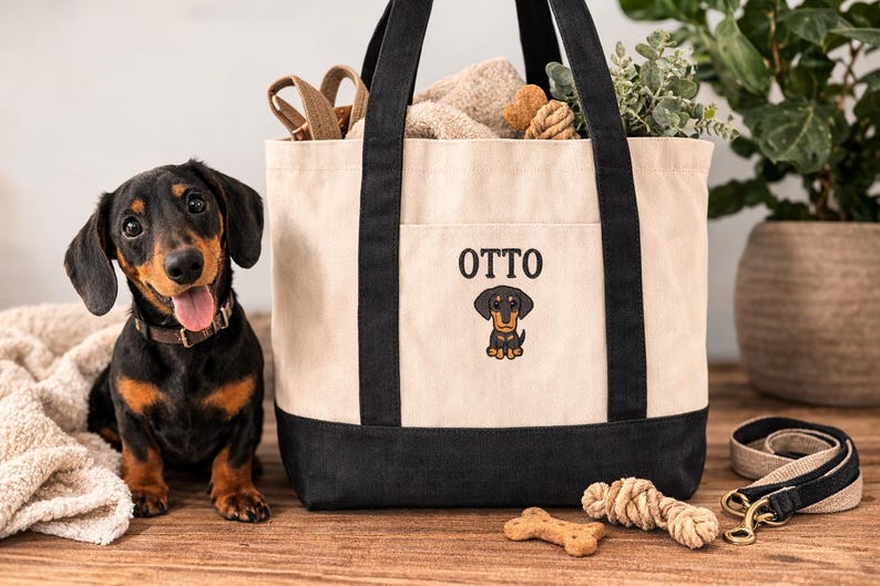 May include: A black and tan dachshund dog sits next to a cream and black tote bag with the name "OTTO" and a dachshund graphic. The bag contains dog toys and is next to a leash and a bone-shaped treat.