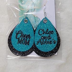 May include: Teal glitter earrings with black lettering. One earring says "Cheer Mom" and the other says "Chloe and Abbie's".