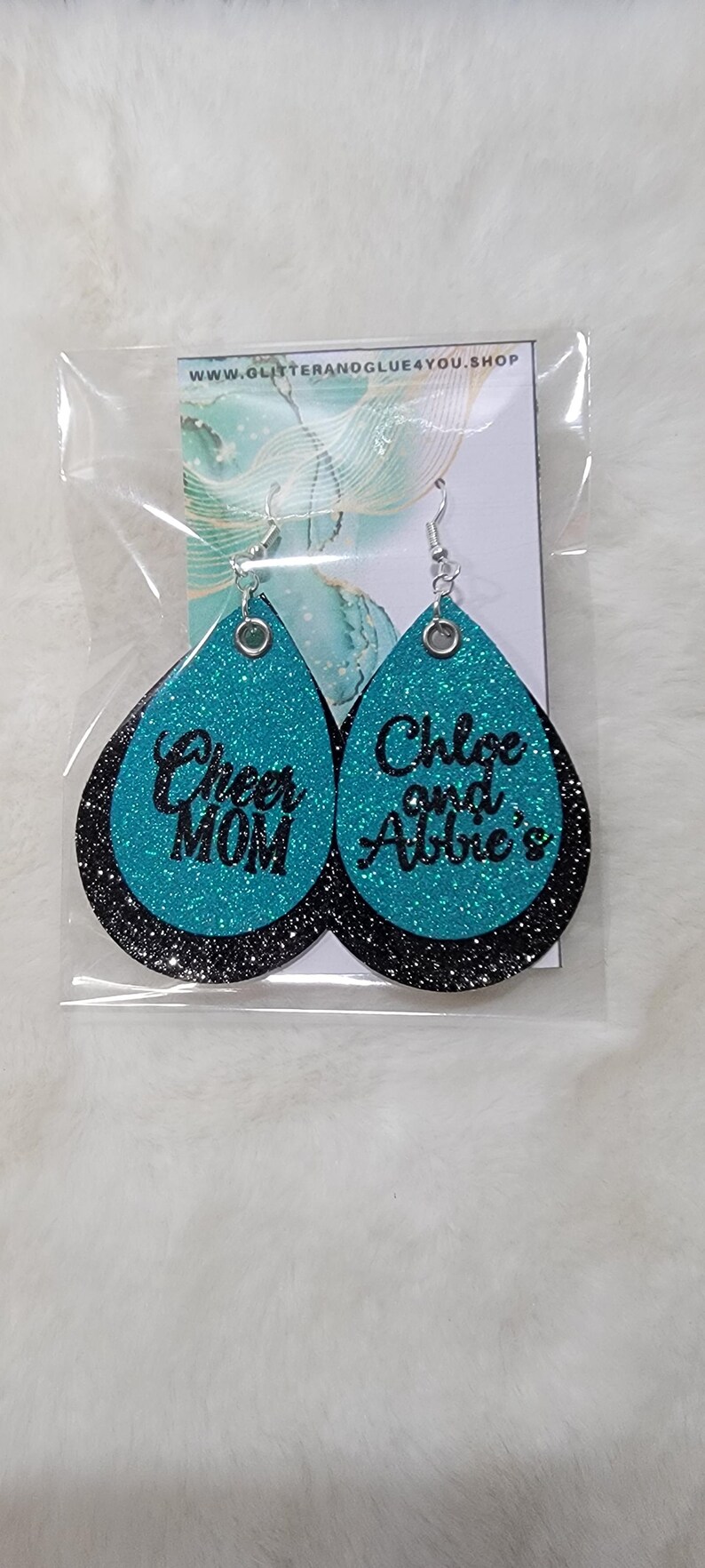 May include: Teal glitter earrings with black lettering. One earring says "Cheer Mom" and the other says "Chloe and Abbie's".